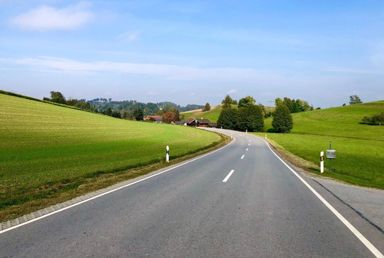 droga podmiejska prowadząca przez pola i łąki i kilka domów wolnostojących obok drogi