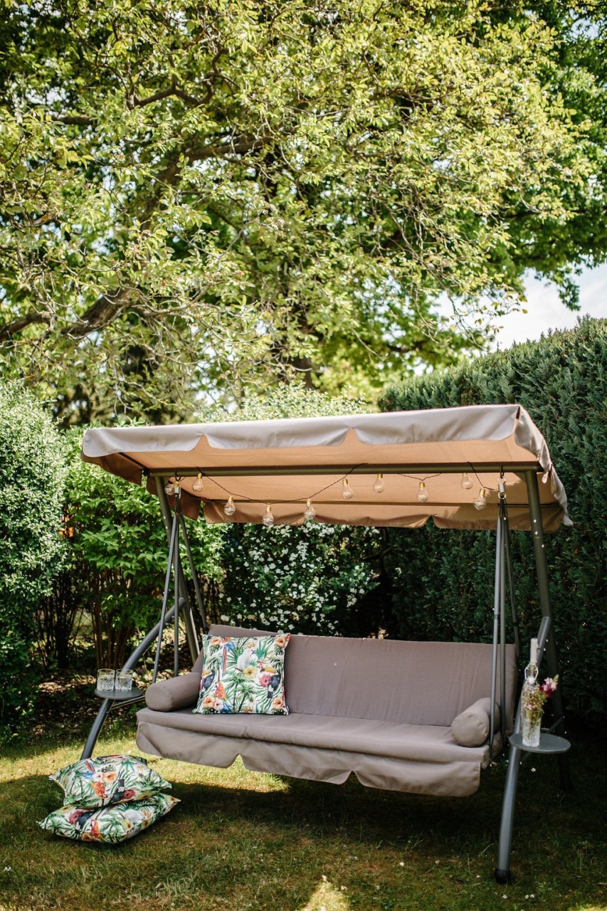 dwuosobowa huśtawka ogrodowa stojąca w ogródku na tle drzew i krzewów